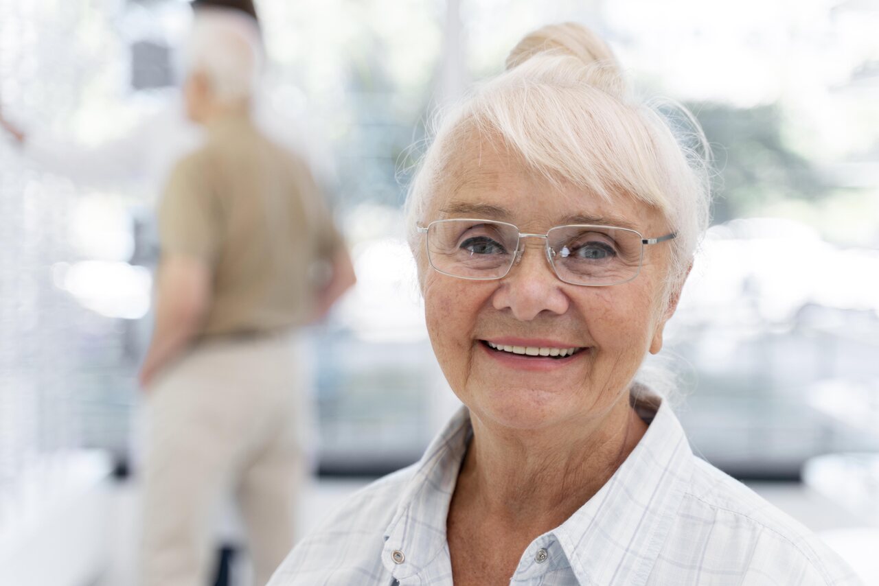 Mulher idosa sorrindo e usando óculos dentro do consultório oftalmológico, fazendo alusão à cirurgia de catarata.