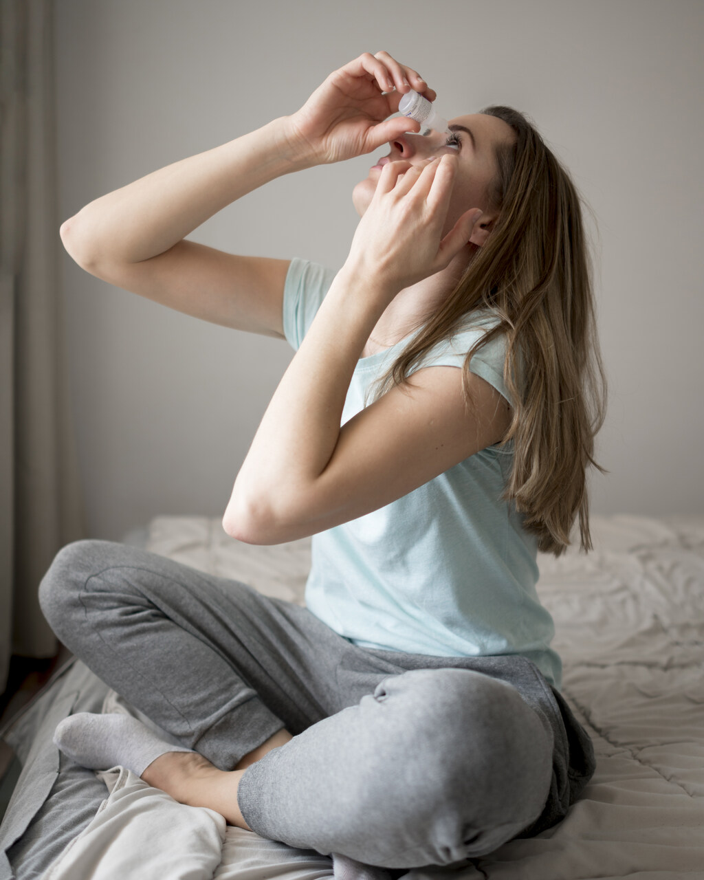 Mulher sentada na cama aplicando colírio nos olhos para ressaltar a relação entre tempo seco e saúde ocular.